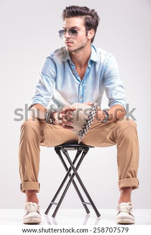Young casual fashion man looking away while sitting on a stool, holding his hat in his hands.