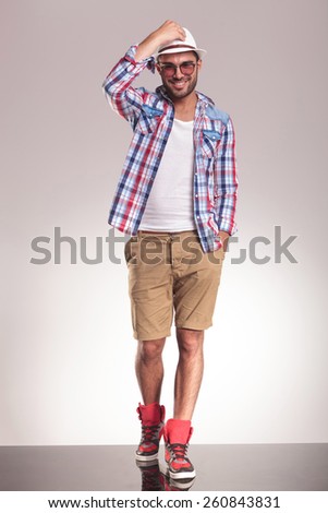 Full body picture of a casual man walking on studio background with one hand in his pocket and the other one on his hat.