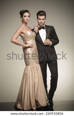 Picture of a young elegant couple posing together, the woman is holding her hand on her waist while the man is fixing his jacket.