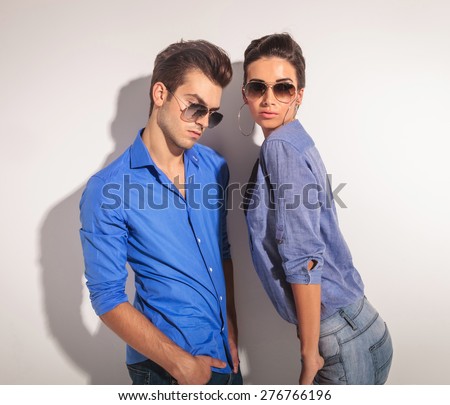 Young fashion couple posing together againt a grey wall. The man is leaning on the wall while looking down and the woman is looking at the camera while holding both hand in the pockets.
