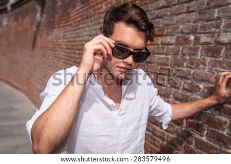 Handsome young man leaning on a brick wall while taking off his sunglasses.
