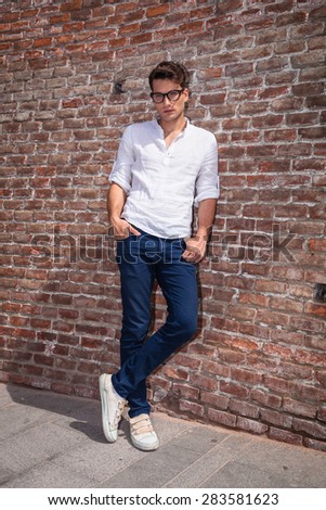 Attractive young man posnig with his hands in pockets while leaning on a brick wall/