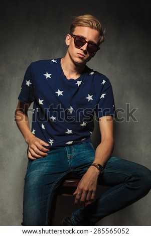 relaxed fashion young man sitting on chair with sunglasses on, in studio