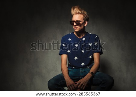 cool fashion model with sunglasses sitting on a stool and looks away from the camera to a side