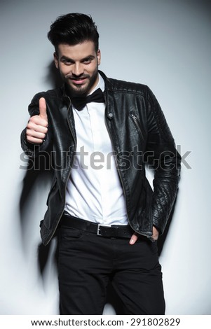 Smiling young business man leaning on a wall with his hand in pocket while showing the thumbs up gesture.