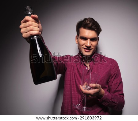 Casual young man presenting a bottle of wine to the camera while holding two glasses in the other hand.