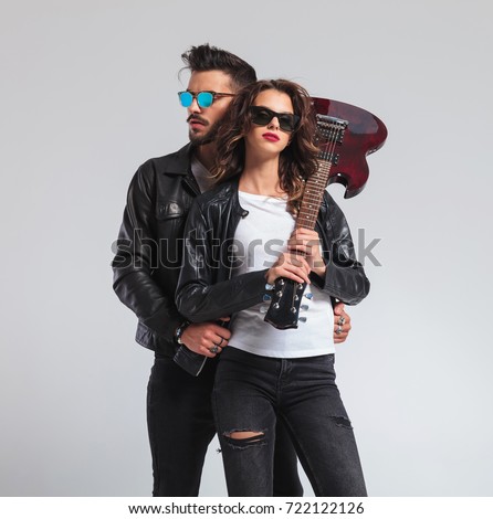 cool woman holding electric guitar on shoulder with man at her back; rock and roll couple posing in studio