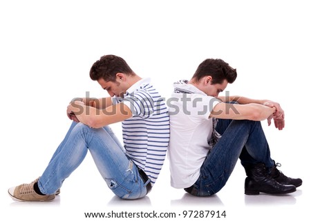 two young casual men sitting back to back on white background and looking very sad
