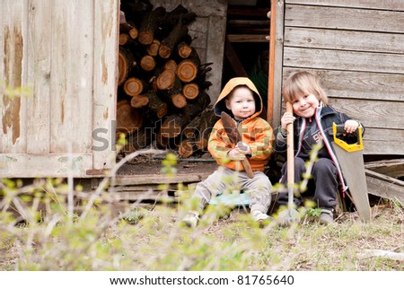 Two little children sit near a shed with garden tools