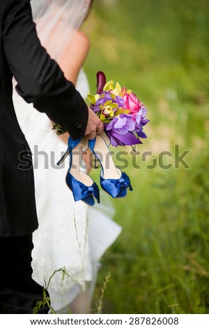 Groom walking behind bride holding beautiful bouquet of flowers and blue shoes