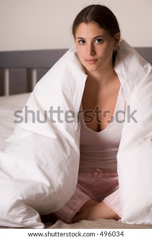 A naturally beautiful young woman sits on her bed with the duvet draped over her shoulders