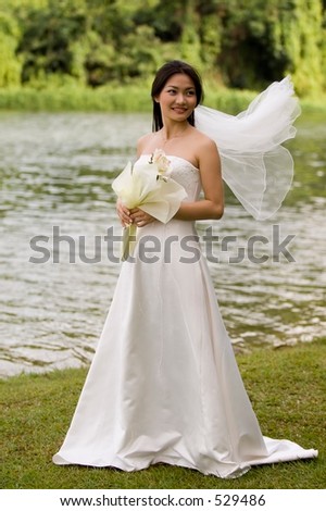 An asian woman stands near a lake in her wedding dress, the veil flying in the wind
