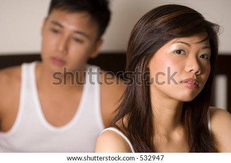 An asian couple sit on a bed, the man behind the woman and out of focus. Serious looking shot.
