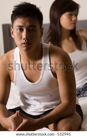 An asian man sits on the edge of the bed with his girlfriend out of focus behind him