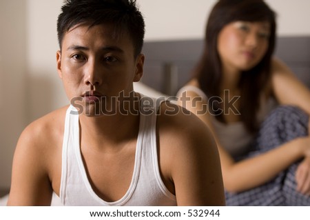 An asian man looks unhappy sitting on the bed, with his partner (out of focus) behind him