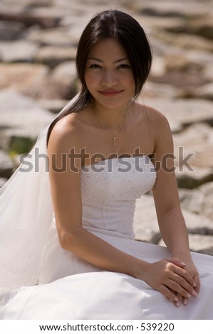 A beautiful asian woman in a wedding dress sits on a rocky beach