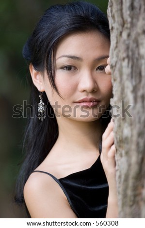 A beautiful young chinese woman looks out from behind a tree, wearing a black evening dress