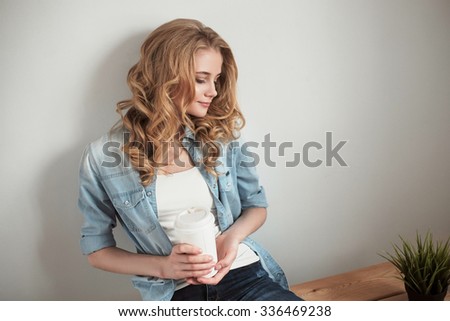 Beautiful young woman with long blonde wavy hair sitting on bench and holding a cup of coffee, looking down