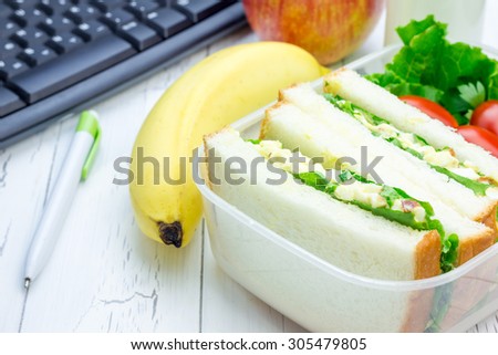 Lunch box with egg salad sandwiches, fruits, and milk on workplace