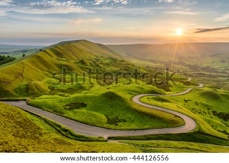 Long and winding rural road leading through green hills in the Peak District, UK at sunset.