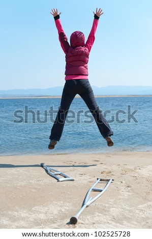 a woman jumps, having left her crutches, at a lake shore, back view