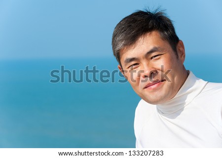 handsome asian young man looks into camera smiling, blue sea and sky