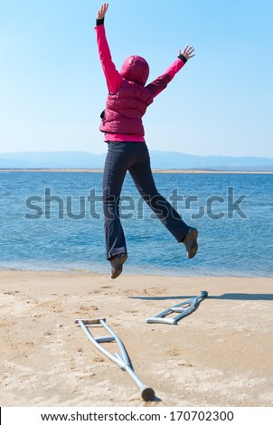 a happy woman jumps, having left her crutches, at a lake shore, back view