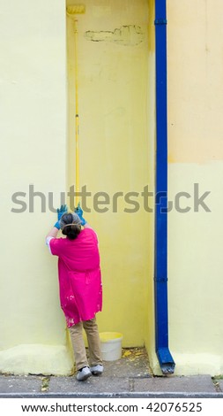 elderly female house painter atiptoe in dark pink overall paints yellow wall with blue downcomer