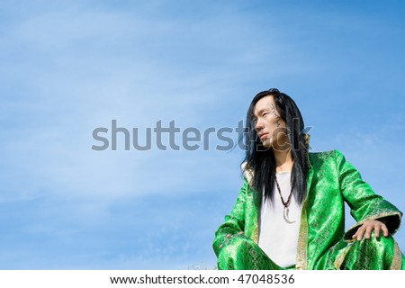 young asian man in green Mongolian suit sits against blue sky, his name Chinghis is written in ancient Mongolian on his cheek, copy space