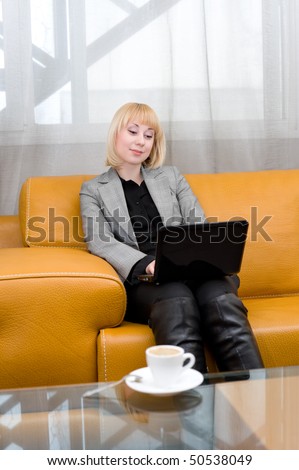 young blonde woman sits on leather sofa with laptop, in front of glass table with cup of coffee