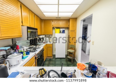 Messy condo kitchen with oak cabinets, tile countertops, gas stove, green flooring and piles of dishes.