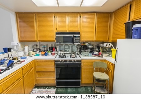 Messy old condo kitchen with oak cabinets, tile countertops, gas stove, green flooring and piles of dishes.