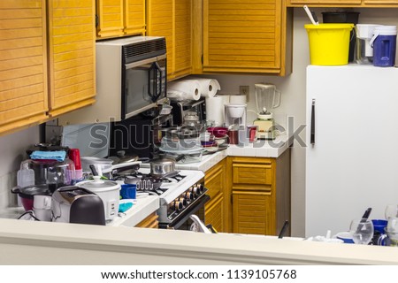 Messy old kitchen with oak cabinets, tile countertops, gas stove, green flooring and piles of dishes.