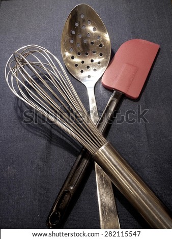 Wire Kitchen Whisk, Slotted Spoon, and Red Colored Spatula on dark Blue Background - Kitchen Essentials in an Isolated Photograph -with Copy Space for your Business Logo, Message, or Advertising Text.