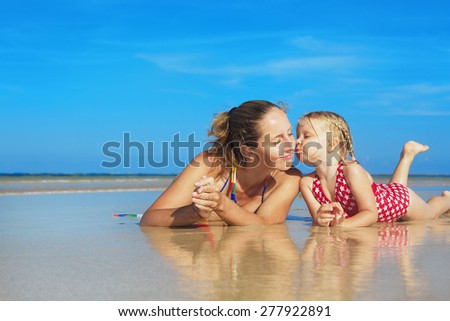 Small child playing with fun and kissing happy young mother smiling with love to her daughter lying on wet sand sea beach. Family lifestyle and summer vacation with children on tropical island resort