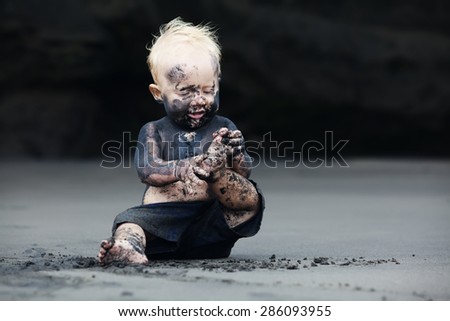 Funny portrait of smiling child with dirty face sitting and playing with fun on black sand sea beach before swimming in ocean. Family active lifestyle, and water leisure on summer vacation with baby