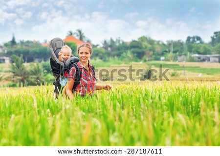 Happy mother travel with baby boy in backpack carrier on green rice terraces. Outdoor activity with child, active and healthy lifestyle during family summer vacation with son on Asian tropical island