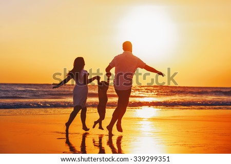 Happy family - father, mother, baby son hold hands and run with fun along edge of sunset sea on black sand beach. Active parents and people outdoor activity on tropical summer vacations with children.