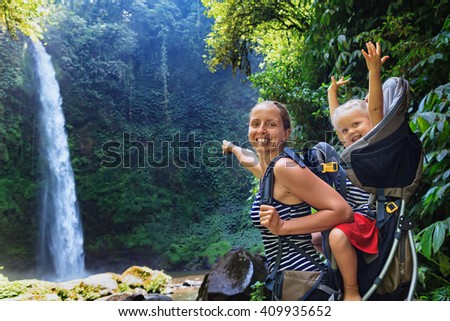 In waterfall pool young happy mother hold little traveller on back - baby girl in carrying backpack enjoying travel adventure, Hiking activity with child on family summer vacation, weekend nature tour