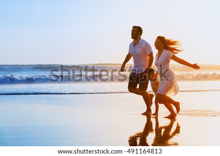 Happy family - father, mother, baby son hold hands and run together with fun along sunset sea surf on black sand beach. Travel, active lifestyle, parents with children on tropical summer vacations.