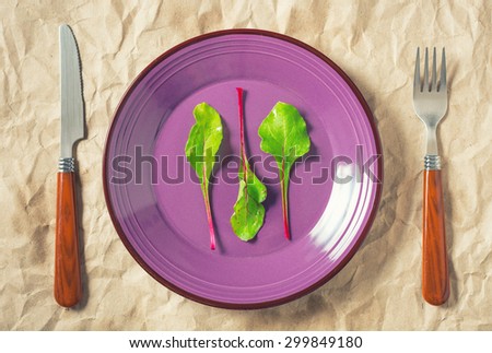 Three beetroot leaves on purple plate with fork and knife on shabby craft paper as a  background. Concept, top view, toned.