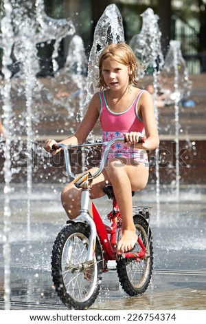 Little girl rides his bike among fountains in summer day