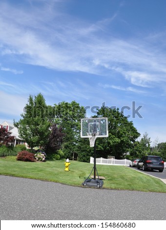 Basketball Hoop on Street Curb of Suburban Neighborhood Home with yellow Fire Hydrant Healthy Lawn Blooming Rhododendrons Parked Cars Sunny blue Sky Clouds USA