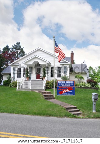 American Flag and Sold real estate sign (another success let us help you buy sell your next home) suburban home residential neighborhood USA blue sky clouds