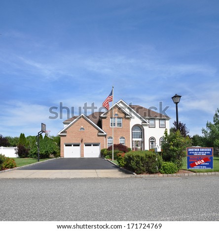 American flag pole Sold Real Estate Sign \'another success let us help you buy sell you next home\' Suburban McMansion style brick home Landscaped sunny residential neighborhood USA blue sky clouds
