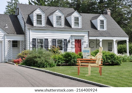 Mannequin sitting on bench reading Real Estate Newspaper how to buy sell a home on front yard lawn of cape code style colonial home in residential neighborhood USA