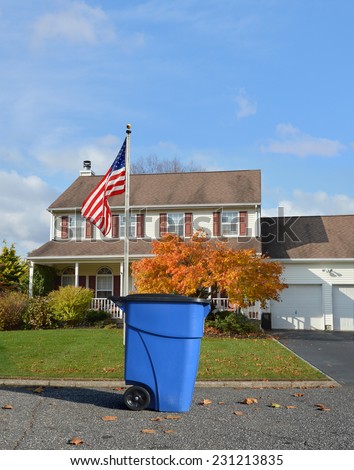 American Flag Pole on front yard lawn Blue Trash Recycle Container on suburban street in front of colonial style home autumn day residential neighborhood blue sky clouds USA