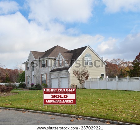 Real Estate sold (another success let us help you buy sell your next home) sign Suburban brick McMansion style home with two car garage white picket fence residential neighborhood blue sky clouds USA