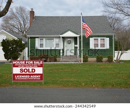 American flag pole real estate sold (another success let us help you buy sell your next home) sign Suburban Bungalow Cape Cod style home autumn day residential neighborhood USA overcast sky