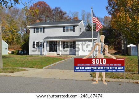 American flag pole Mannequin holding Real estate sold (another success let us help you buy sell your next home) sign Suburban Gray High Ranch home autumn day residential neighborhood USA